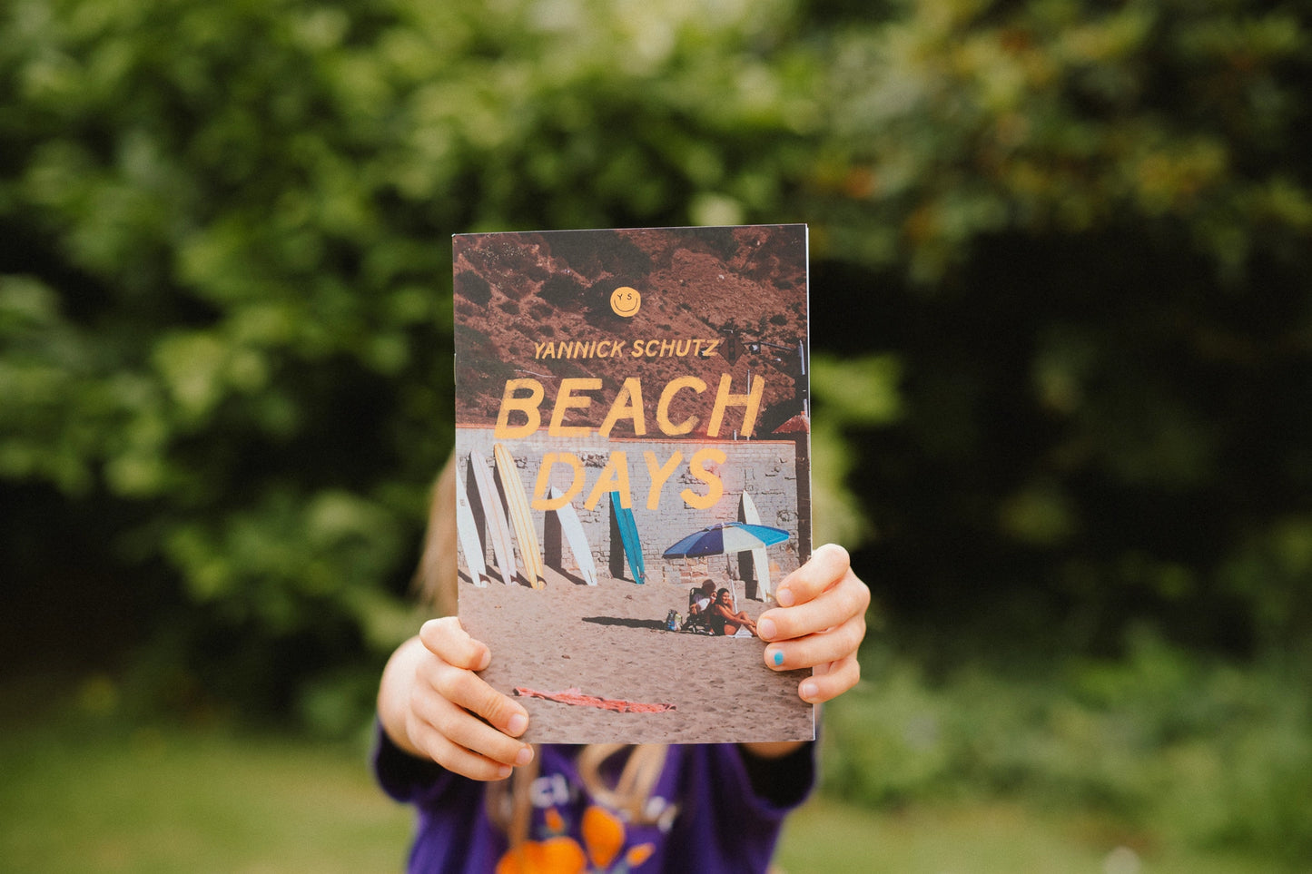 Person holding a book titled 'Beach Days' by Yannick Schutz in front of a blurred green background