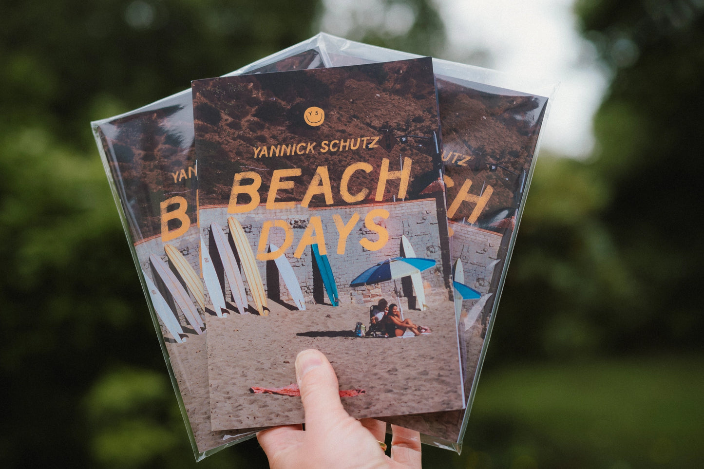 Three 'Beach Days' books by Yannick Schutz held in a hand with a blurred natural background